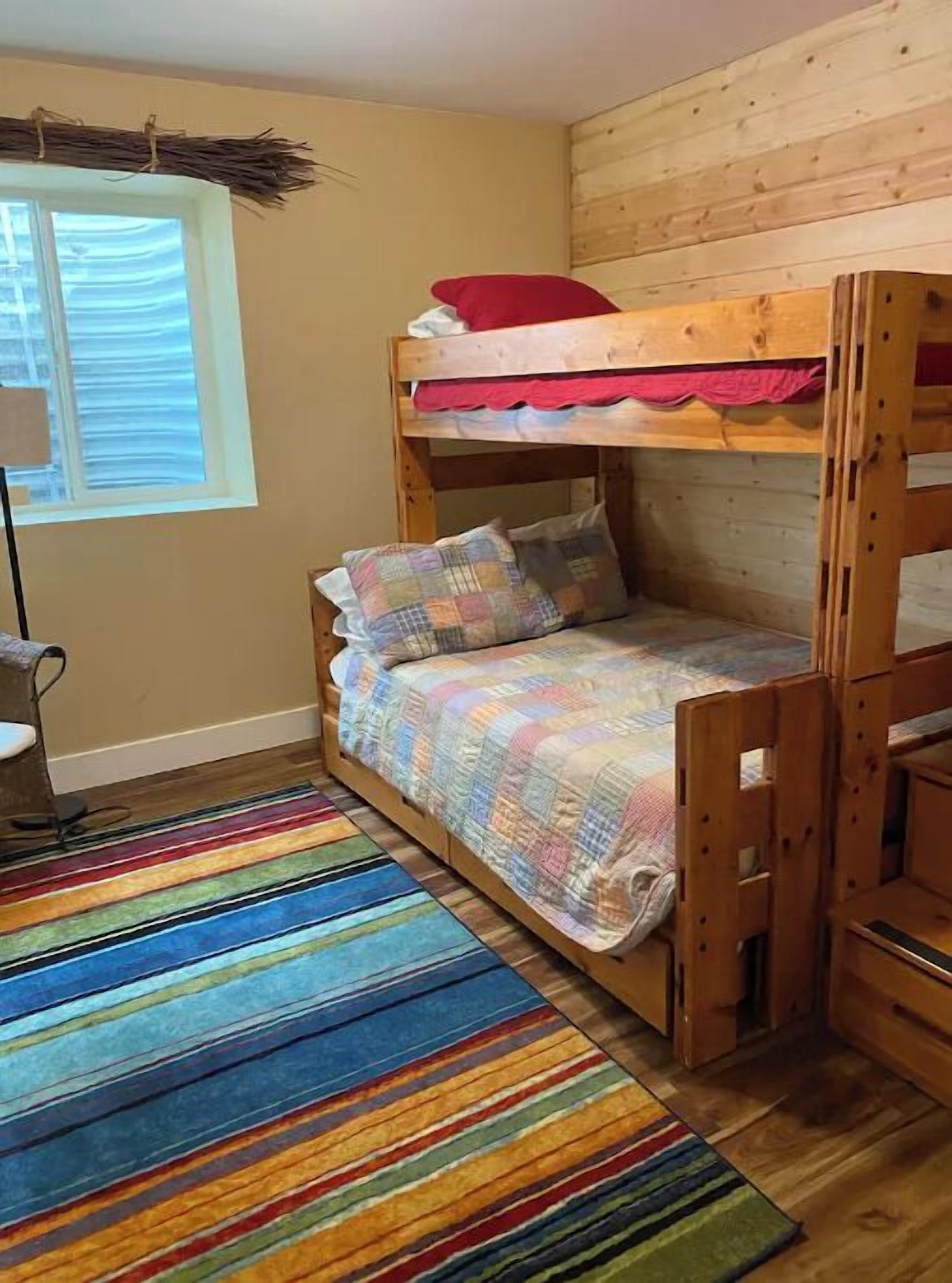 A bunk bed room with a colorful striped rug, wooden walls, and a window with blinds.