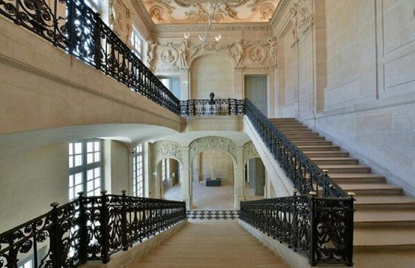 Uma escadaria imponente com grades pretas ricamente decoradas e piso xadrez, que leva a um nível superior com portas em arco e teto ornamental.