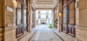 Um corredor estreito com portas e colunas de madeira decorada, que leva a um edifício com várias janelas.