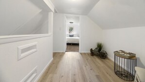 A narrow hallway with wooden flooring, white walls, and a ceiling light fixture. A bed is visible through an open doorway at the end of the hallway.