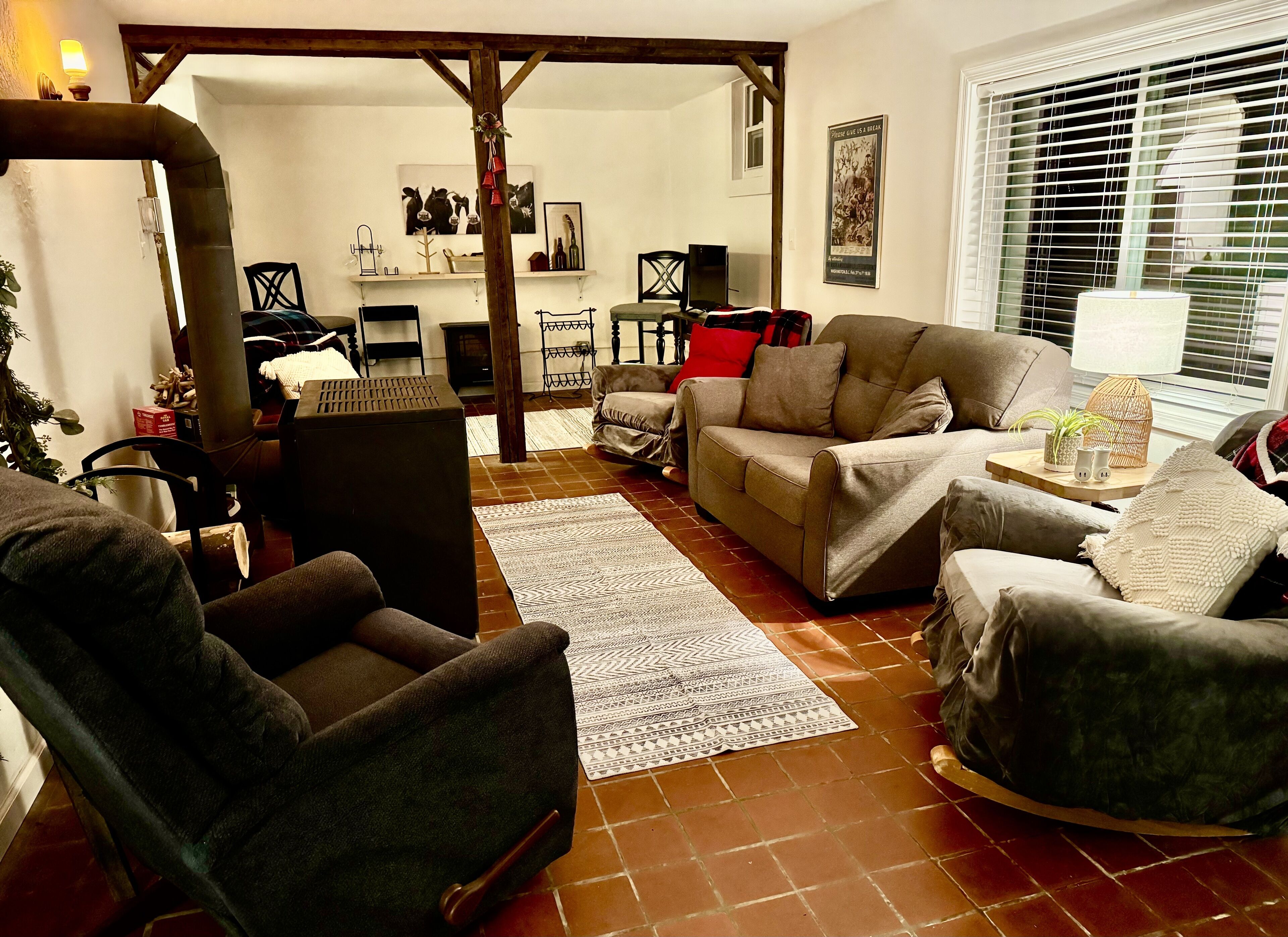 A cozy living room with a brown sofa, a dark armchair, and a patterned rug. There is a wooden beam structure, a window with blinds, and a wall-mounted shelf with decorative items.