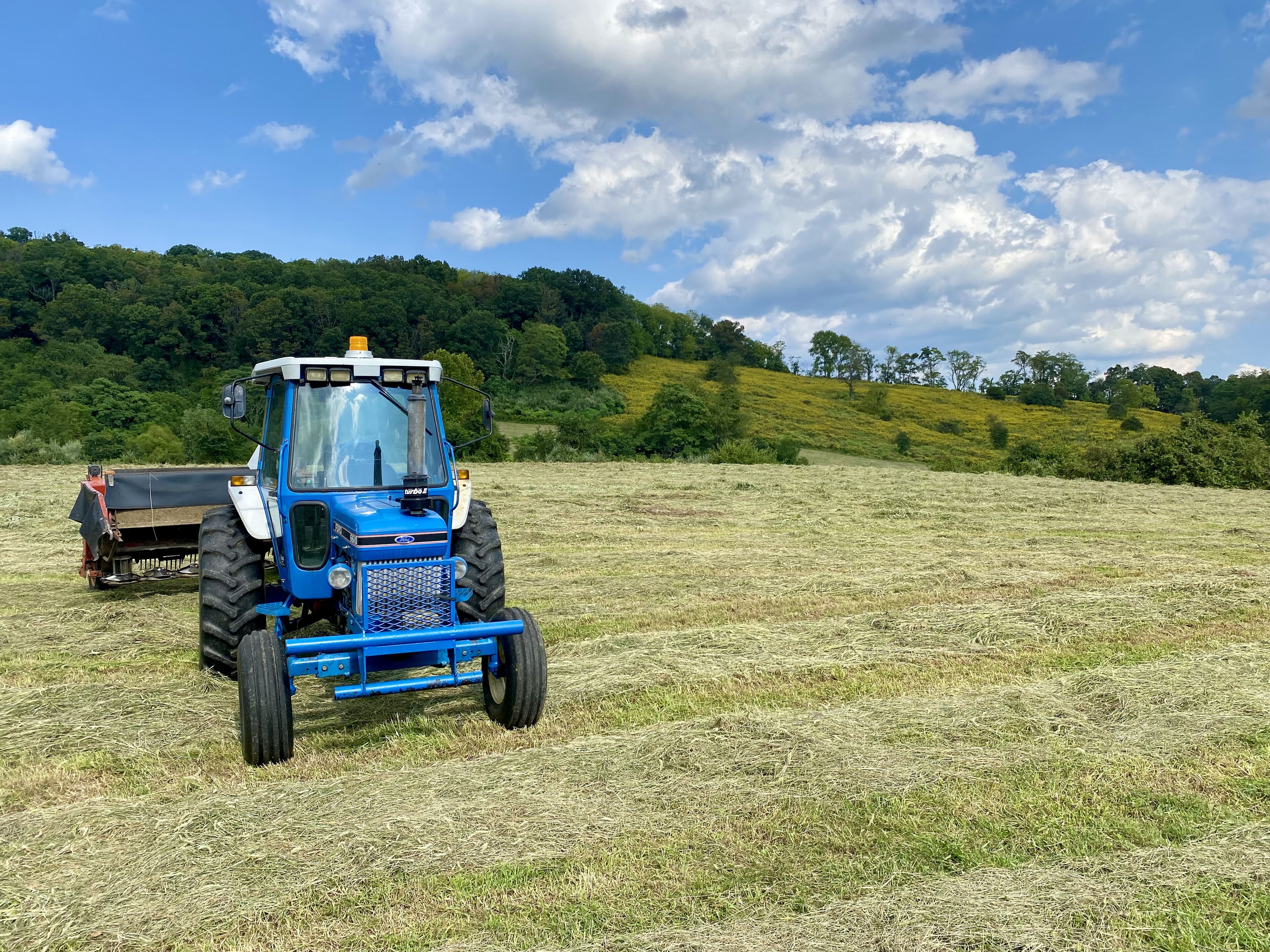 Un tractor blau en un camp amb un remolc, envoltat d'unes verdes muntanyes i un cel amb nuvolades.