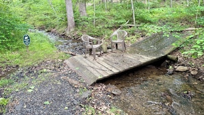 Eine Holzbrücke über einen Bach, auf der linken Seite zwei Plastikstühle.