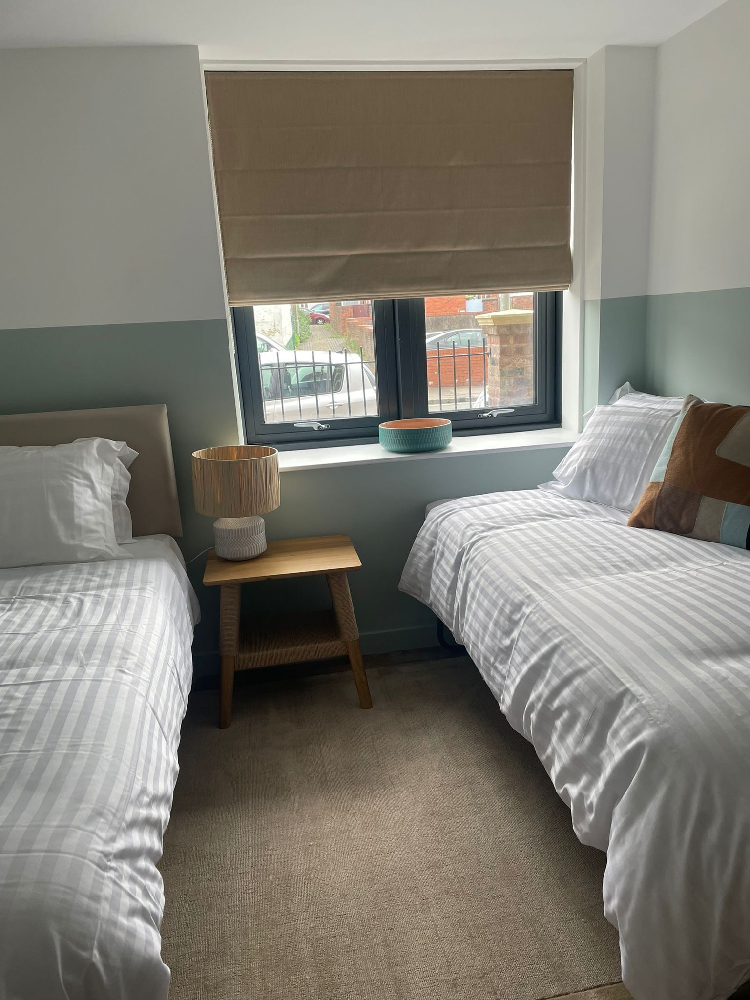 Ein Schlafzimmer mit zwei Betten, einem Fenster mit Blick auf die Straße, einem Nachttisch mit Lampe und einem Fensterrollo.
