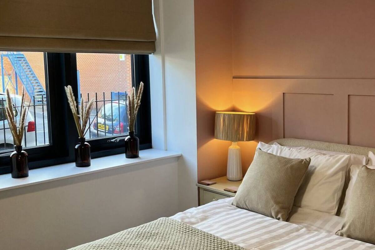 Une chambre d’hôtel avec un grand lit, une fenêtre donnant sur un immeuble et une échelle de secours bleue, une table de chevet avec une lampe, et deux vases contenant des plantes séchées sur le rebord de la fenêtre.