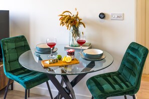 Une table ronde en verre, accompagnée de deux chaises à revêtement vert, un vase contenant des fleurs séchées, deux verres à vin, un bol et une planche en bois sur laquelle sont disposés du fromage et un citron.