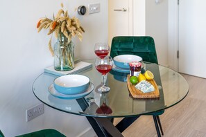 Una mesa de vidrio con platos azules, dos copas de vino, un tarro de mermelada y una tabla de madera con queso y fruta.