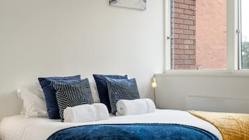 Un dormitorio con cama, almohadas, una colcha, una obra de arte en la pared y una ventana con vista a un edificio de ladrillo.