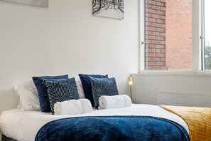 Un dormitorio con cama, almohadas, una colcha, una obra de arte en la pared y una ventana con vista a un edificio de ladrillo.