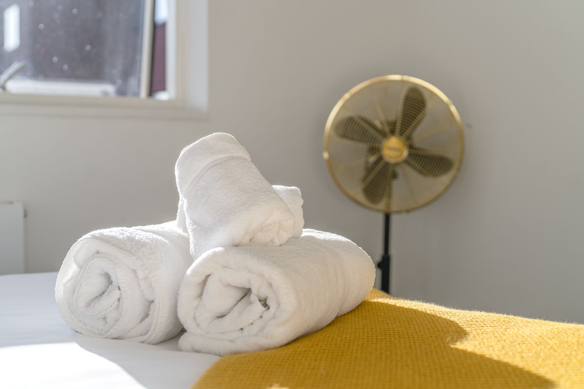 A neatly folded white towel on a bed with a yellow blanket, a standing fan, and a window in the background.