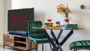 Un salón moderno con un letrero de Netflix, un sillón de terciopelo verde, una mesa de vidrio con una copa de vino y un jarrón con plantas secas.