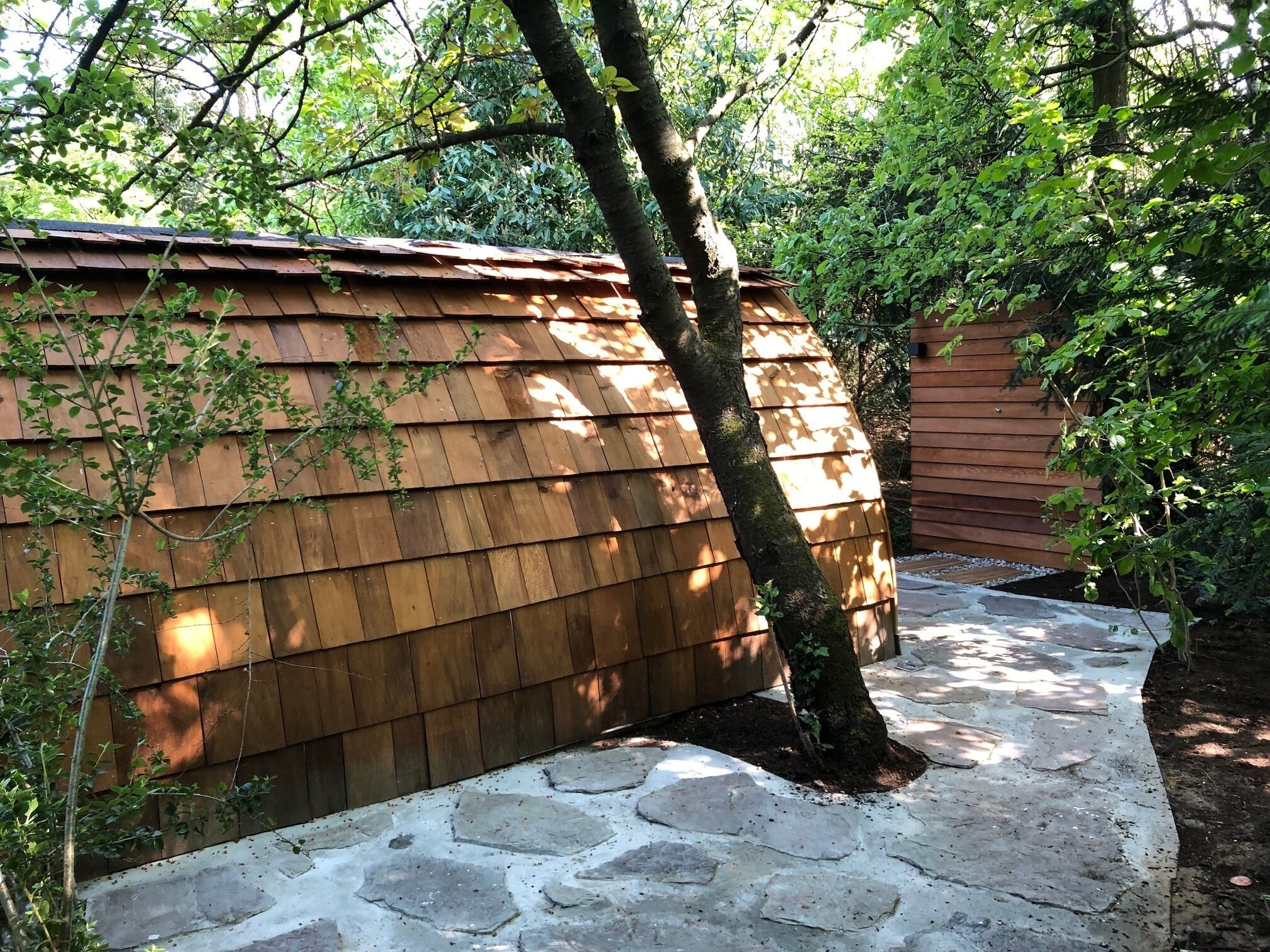 A wooden shed with a stone pathway leading to it, surrounded by trees and greenery.