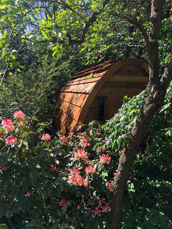 A rustic wooden structure with a sloped roof, surrounded by lush greenery and blooming pink flowers.