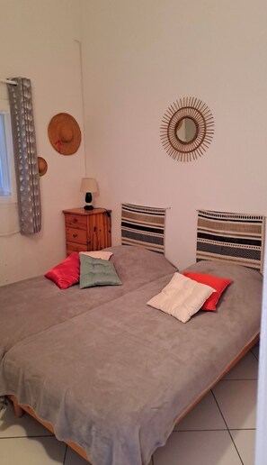 A bed with a grey bedspread, two pillows, a wooden nightstand with a lamp, a wall mirror, and a window with curtains. - holiday home in residential area near the sea (Port-la-Nouvelle)