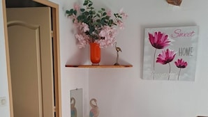 Een kamer met een blauwe stoel, een houten deur, een klein plankje met een bloevas, een kunstwerk aan de muur en een betegelde vloer.