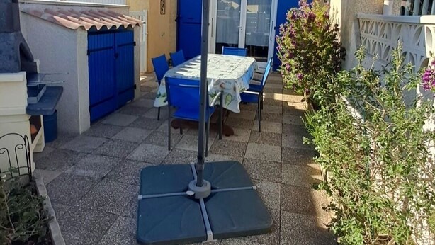 Een terras met een tafel, stoelen, een blauwe parasol en een barbecue.