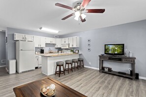 Eine moderne Küche mit weißen Schränken, einer zentralen Kochinsel mit Barhocker, einem Deckenventilator und einem Fernseher auf einem Holzbodenständer.