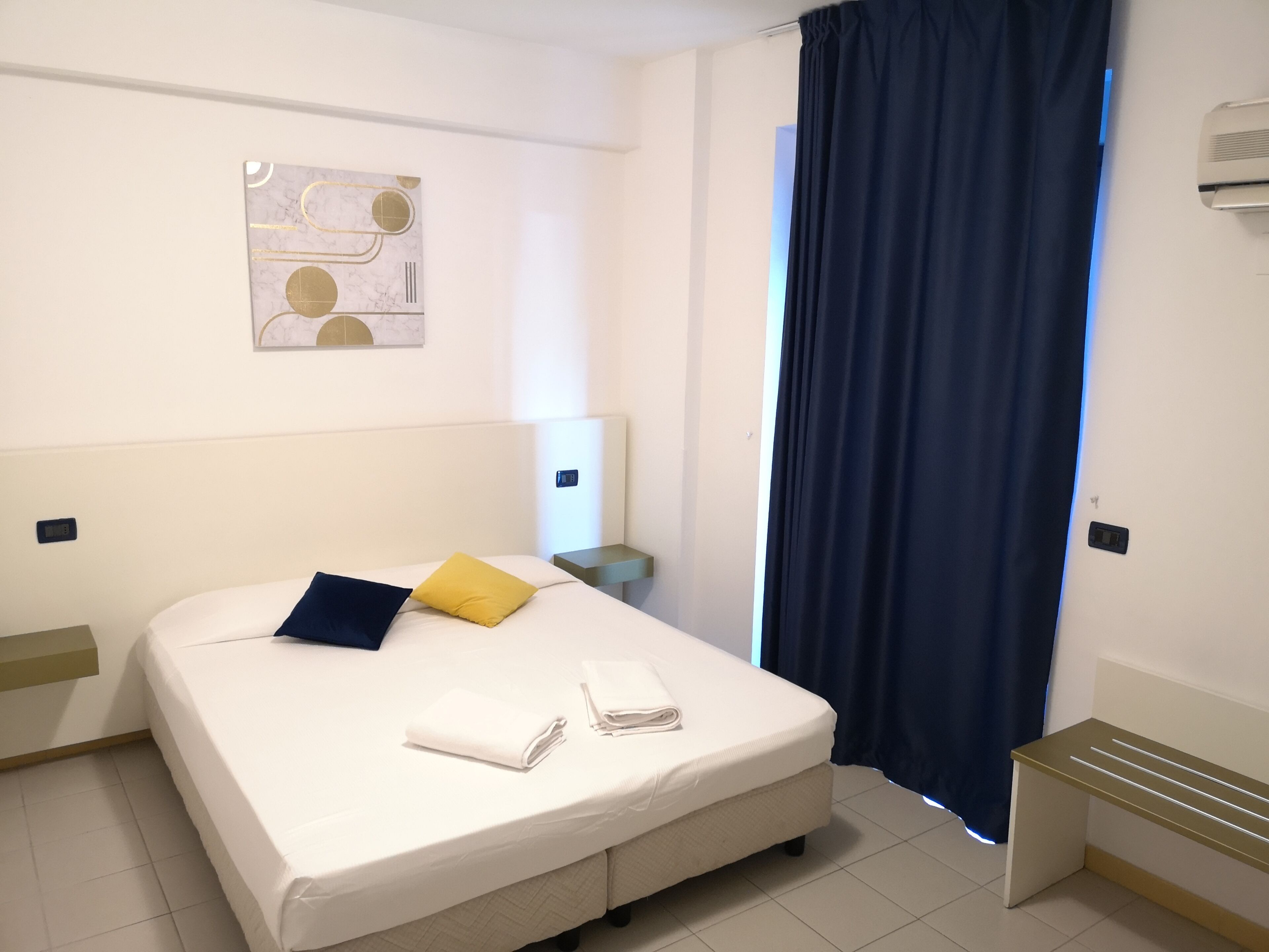 A neatly made bed with pillows, a bedside table, a wall-mounted air conditioner, and a window with a curtain.