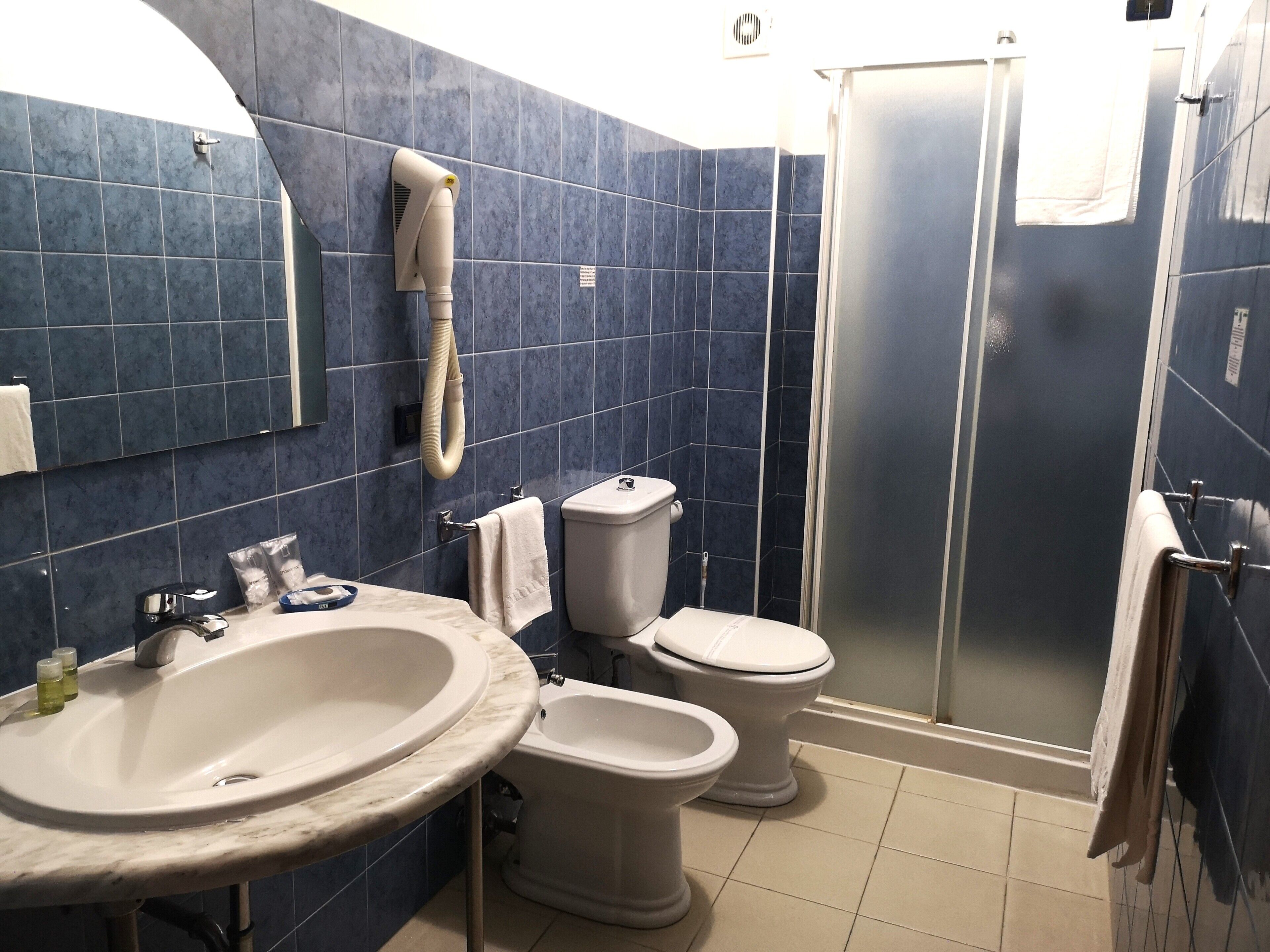 Un baño con lavamanos, dos inodoros, ducha y secador de pelo fijado a la pared.