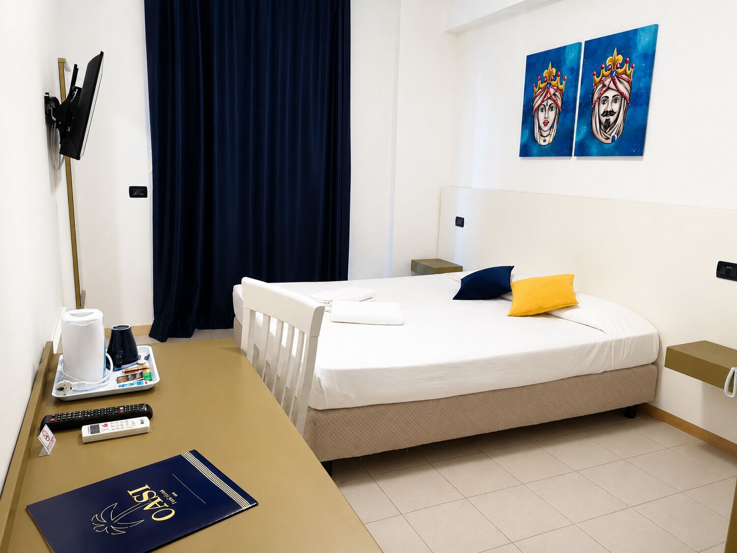 Una camera d'albergo con un letto, una scrivania con vassoi per la colazione, un telecomando, un libro e opere d'arte alle pareti.