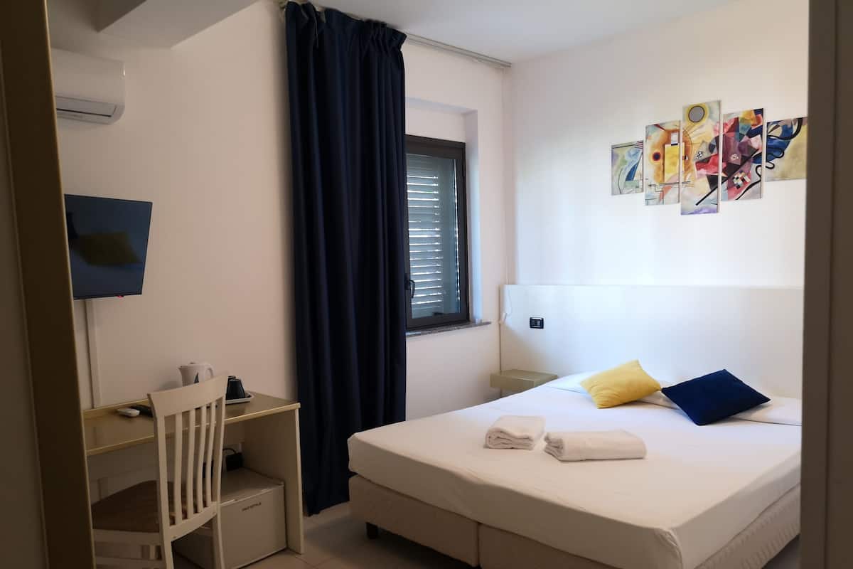 Quarto de hotel com cama, escrivaninha, cadeira, janela com persianas e uma parede com obras de arte.