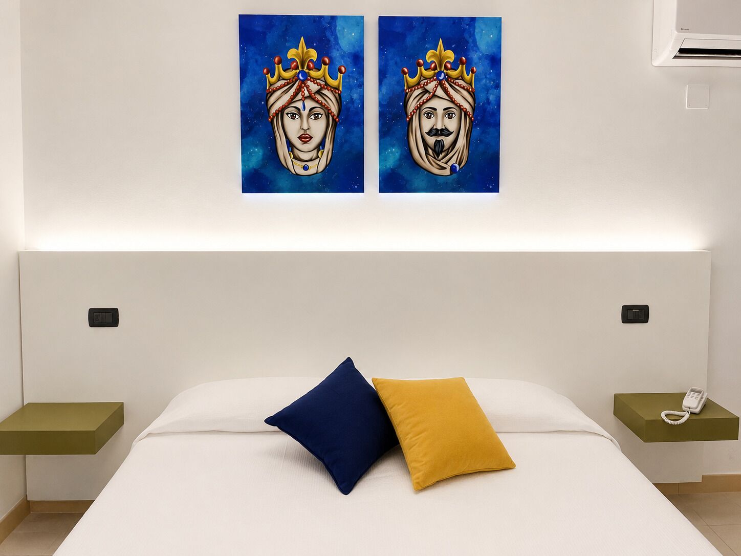 Hotellihuone, jossa on sänky, kaksi yöpöytää, toisessa yöpöydässä puhelin ja seinällä kaksi taulua.
