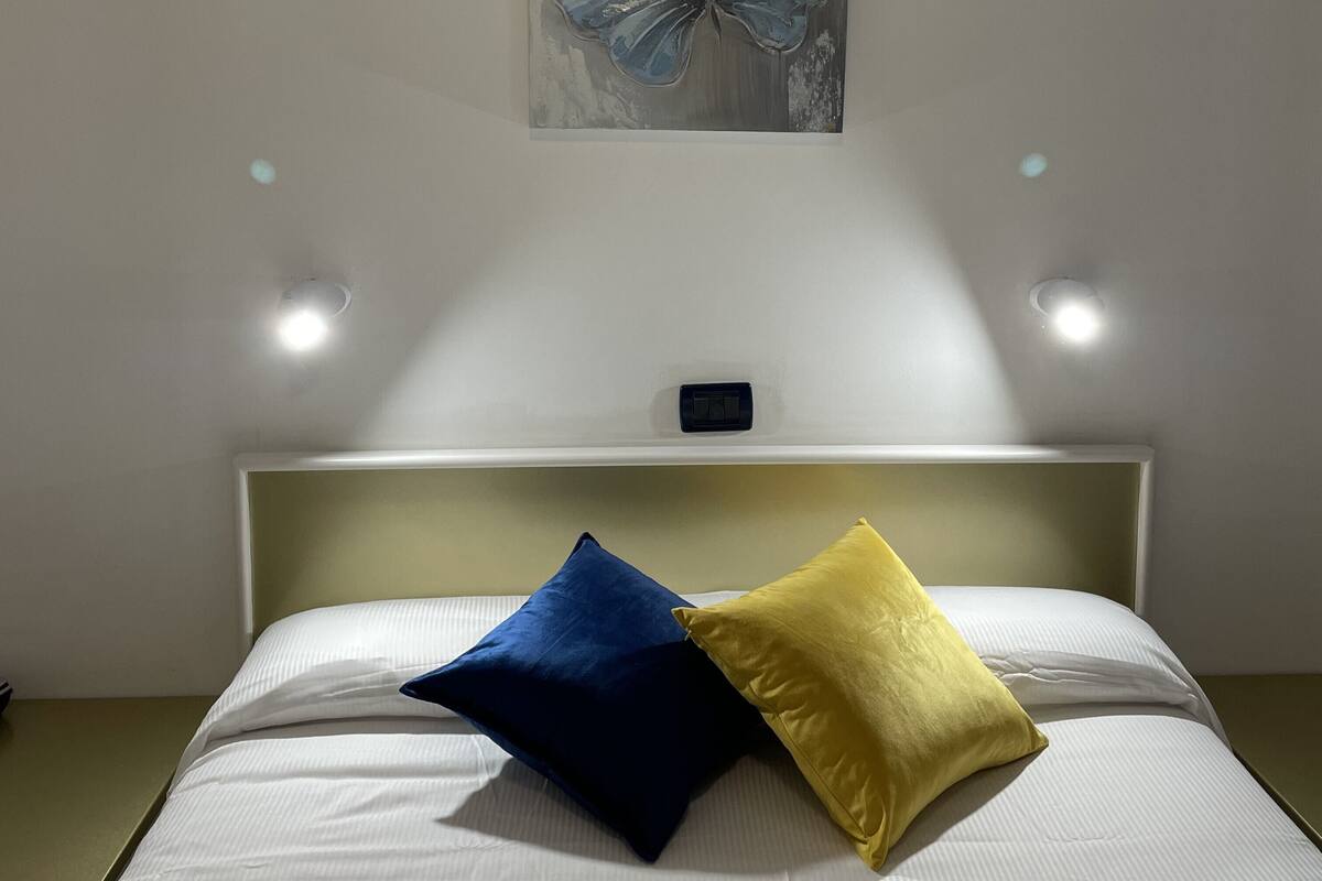 Cama bem arrumada com duas almofadas, interruptor de luz na parede e um quadro sobre a cama.