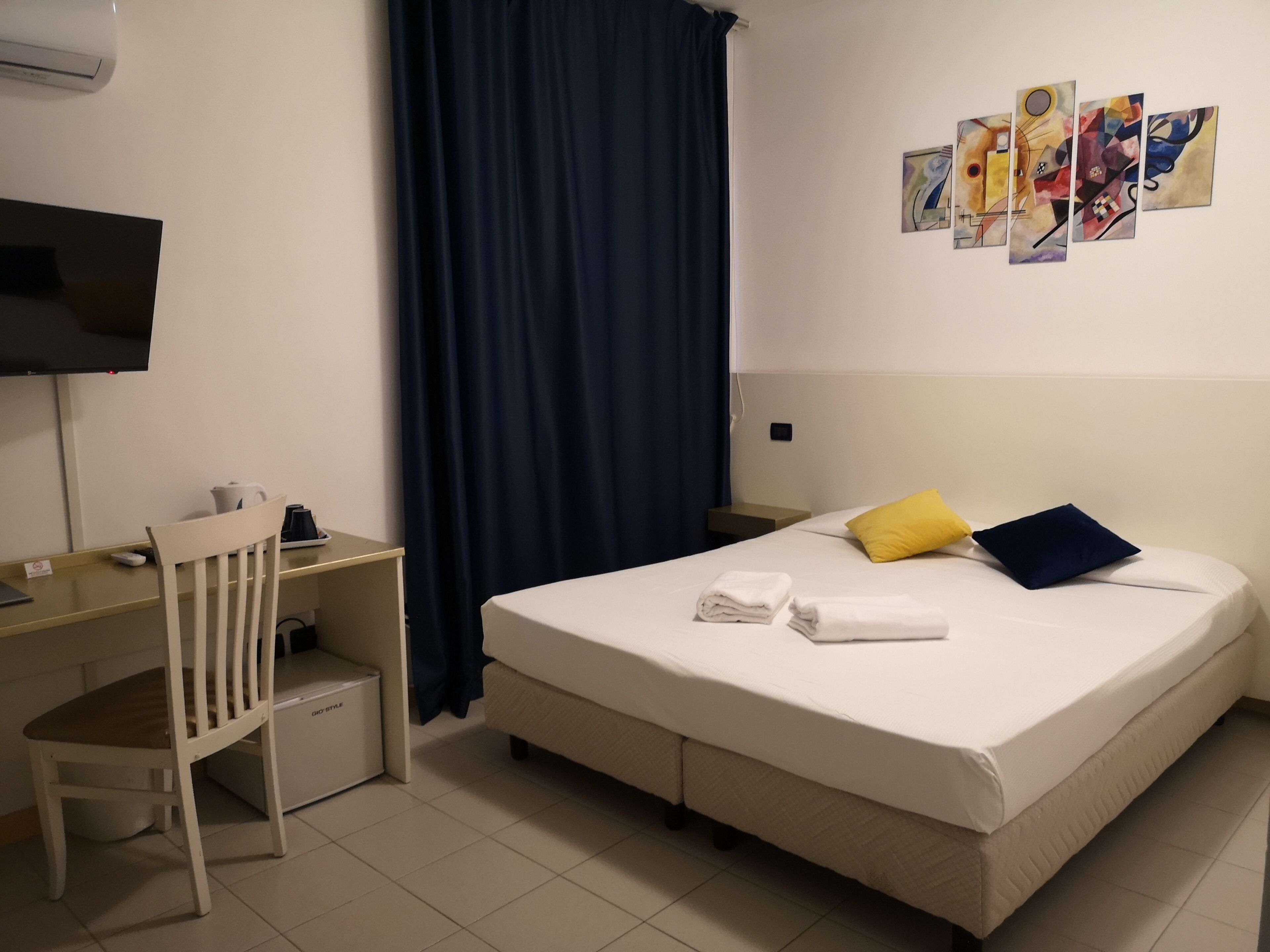 A hotel room with a bed, a desk, a chair, a television, and artwork on the wall.