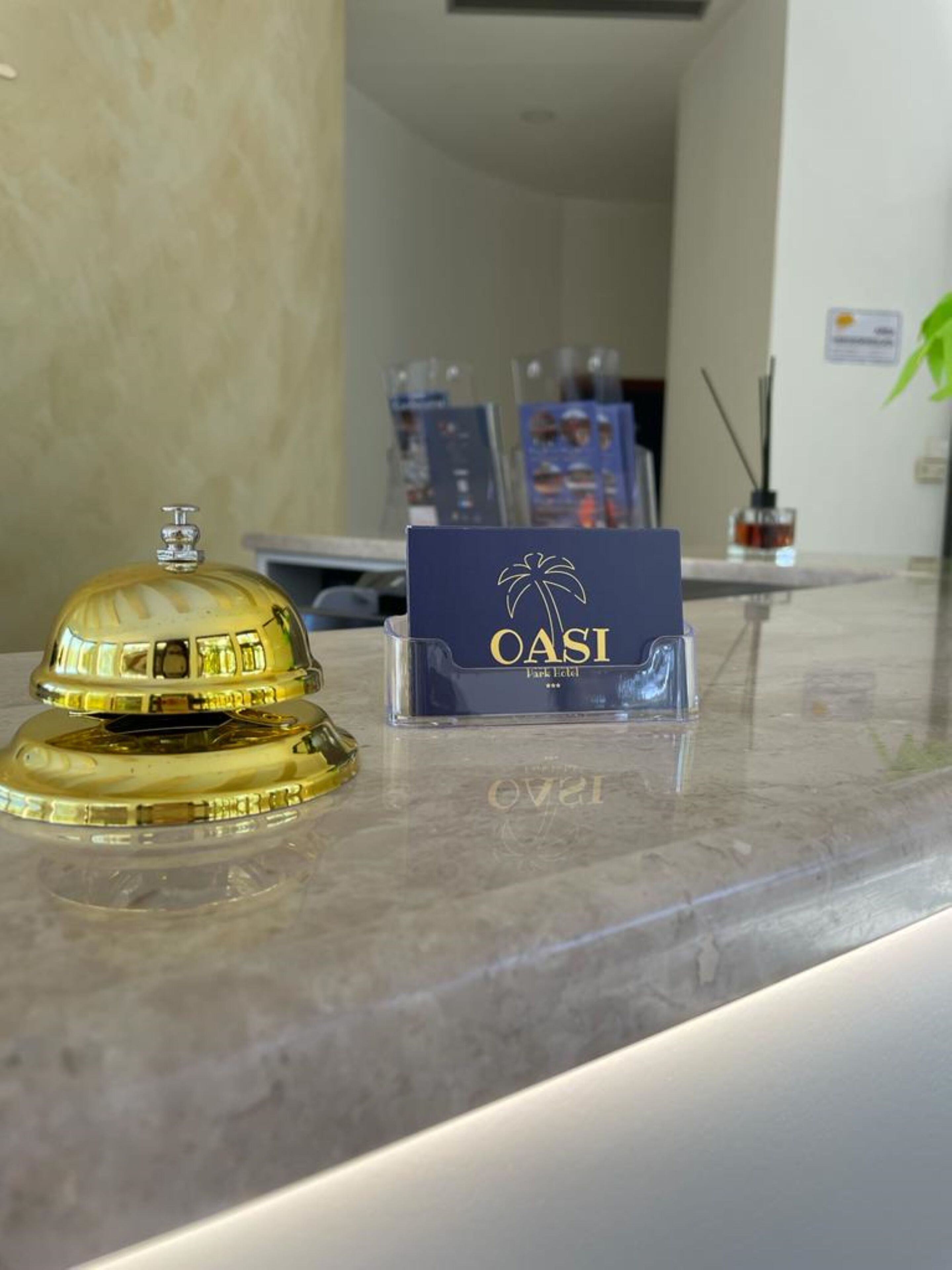 A reception desk with a golden bell, a blue OASI Park Hotel sign, and a small bottle of hand sanitizer.