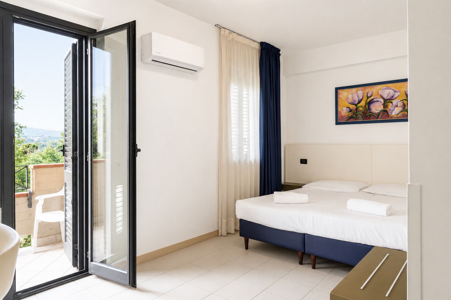 A hotel room with a bed, a painting, an air conditioning unit, a sliding glass door, and a view of greenery.