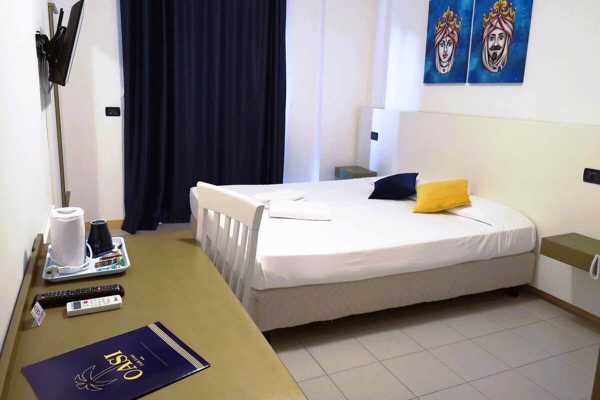 Quarto de hotel com uma cama, escrivaninha, laptop, controle remoto, um livro e uma cortina.