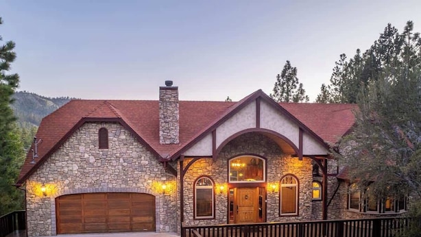 A stone house with a red roof, a wooden garage door, and a stone chimney. - Canyon Crest Lodge. Hot Tub, Game Room, and Views. (Big Bear Lake)
