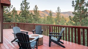 A deck with Adirondack chairs and a fire pit, overlooking a forested mountain range. - Canyon Crest Lodge. Hot Tub, Game Room, and Views. (Big Bear Lake)