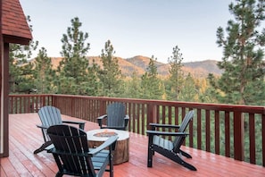 A deck with Adirondack chairs and a fire pit, overlooking a forested mountain range.