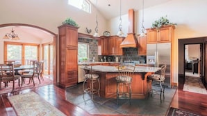 A spacious kitchen with wooden cabinets, a large island with bar stools, and a hanging light fixture. - Canyon Crest Lodge. Hot Tub, Game Room, and Views. (Big Bear Lake)