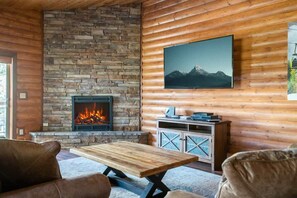 A log cabin interior with a stone fireplace, wooden walls, a mounted TV, and a wooden table.