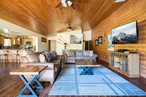 A spacious living area with a wooden ceiling, a sofa set, a coffee table, and a television mounted on the wall.