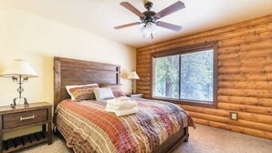 Quarto com parede de madeira rústica, cama com colcha estampada, duas luminárias de cabeceira, mesa de cabeceira com gavetas, ventoinha no teto e janela com persianas.