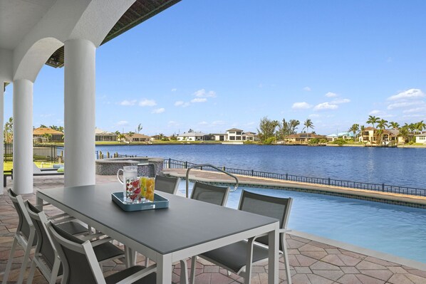 Un espace aménagé au bord de la piscine, avec une table et des chaises, une carafe de fruits et une vue sur un lac, avec des bâtiments en arrière-plan.