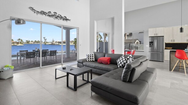Una sala moderna con un sofá gris, cojines negros y blancos, y una almohada roja. Cuenta con un comedor con una mesa y sillas, y una cocina con refrigerador y armarios.