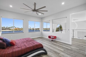 Une chambre moderne avec un lit, de grandes fenêtres, un ventilateur de plafond et une vue sur une étendue d’eau.