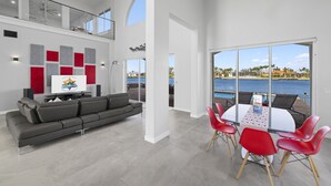 Un salón amplio con un sofá gris, una mesa de comedor con sillas rojas y un gran ventanal con vista a un cuerpo de agua.