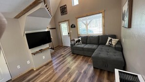 Un salón estilo loft con un sofá seccional gris grande, un televisor de pantalla plana montado en una viga de madera y una escalera que conduce a un nivel superior.