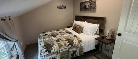 A bedroom with a bed, a nightstand, a lamp, a window with curtains, and a door.