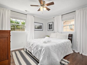 A bedroom with a bed, white curtains, a ceiling fan, and framed pictures on the wall.