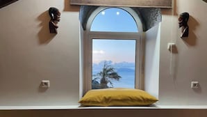 A room with a window overlooking a palm tree and mountains, a yellow pillow on a ledge, and two wooden head sculptures on the walls.