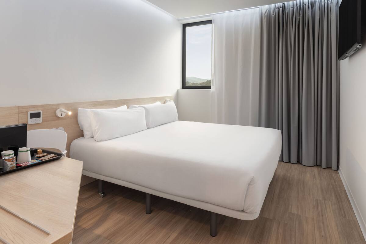 Una habitación de hotel moderna con una cama bien hecha, una mesita de noche con un bandeja de artículos de tocador, un televisor de pantalla plana y una ventana con cortinas.