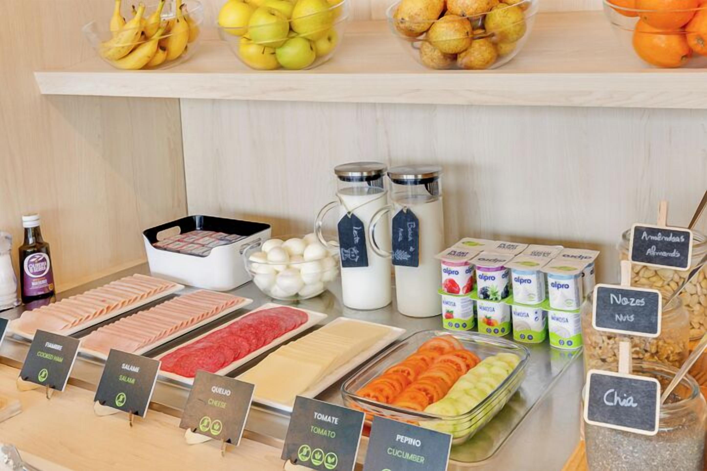 Exhibición de alimentos con diferentes productos, incluyendo jamón cocido, salami, queso, tomate, pepino, y chía. También hay frascos con frutos secos y productos lácteos.
