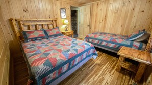 Ein Zimmer mit Holzwänden, ausgestattet mit zwei Betten, jedes mit einer gesteppten Bettdecke. Auf der einen Seite befindet sich ein Nachttisch mit Lampe, auf der anderen eine kleine Holztisch.
