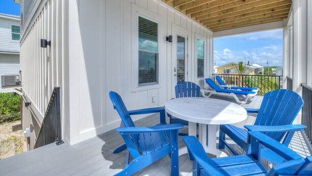 Un patio techado al aire libre con una mesa redonda y sillas Adirondack azules, con vista a una zona residencial.